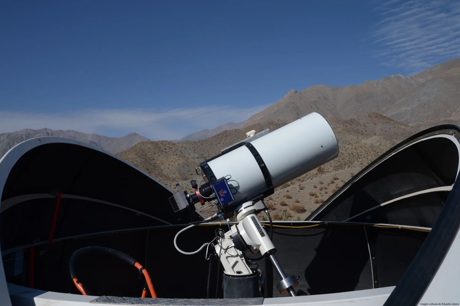 A telescope on its mount under an open dome shutter, with dry mountains behind